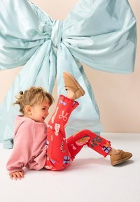 Leggings rouges à motifs festifs, sweat-shirt rose oversize et chaussures marron à enfiler. En arrière-plan, un grand nœud bleu clair.