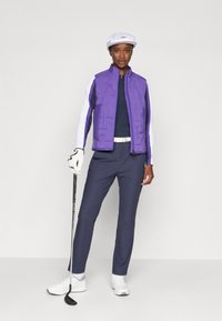 Gilet isolant violet sur un haut bleu foncé, pantalon marine et baskets blanches. Tient un club de golf, portant une casquette blanche et un gant de golf.