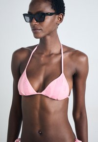 Rosa Bikini-Oberteil mit dreieckigem Design, dünnen Trägern und glatter Textur. Modell trägt schwarze Sonnenbrille und steht vor einem neutralen Hintergrund.