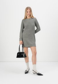 Szürke bordázott kötött ruha hosszú ujjakkal, fekete táska, fehér zoknik és hegyes fekete magassarkúak, minimalista környezetben megjelenítve.