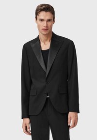 Uomo che indossa una giacca da smoking nera con lapels in satin e pantaloni neri, in piedi davanti a uno sfondo grigio chiaro uniforme.