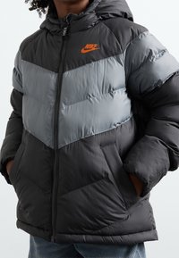 Svart och grå vadderad jacka med chevrondesign, dragkedja, huva och orange Nike-logotyp. Har quiltad struktur och sidofickor.