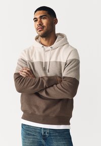 Odranžirana jopa s kapuco sledi vodoravnim bež, sivim in rjavim črtam. Izdelana je iz mehke tkanine z rebrastim robom in zavezovanjem s špagetom.