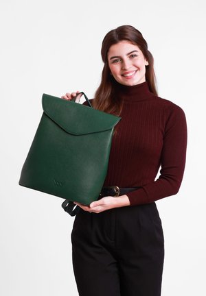 Tmavě zelený kožený batoh s klopou, texturovaným povrchem a nastavitelnými popruhy, který drží osoba oblečená v vínovém roláku.