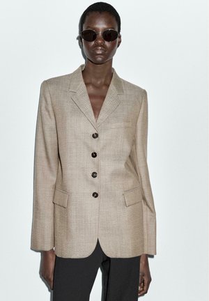 Beige herringbone blazer with notched lapels, four brown buttons, and two front pockets, worn over black trousers. Round sunglasses complete the look.