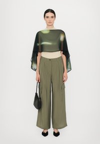 Läskig svart kappa över en beige skjorta, parat med lösa olivgröna byxor med sidofickor, och svarta spetsiga ballerinas tillsammans med en vävd väska.