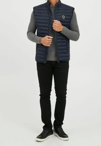 Navy insulated vest with quilted texture, high collar, zipper front, and logo detail. Paired with a grey long-sleeve shirt and black pants.