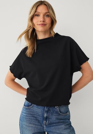Vrouw met blond haar die een losse zwarte top en blauwe jeans draagt, staat met handen in de zij tegen een effen lichte achtergrond.