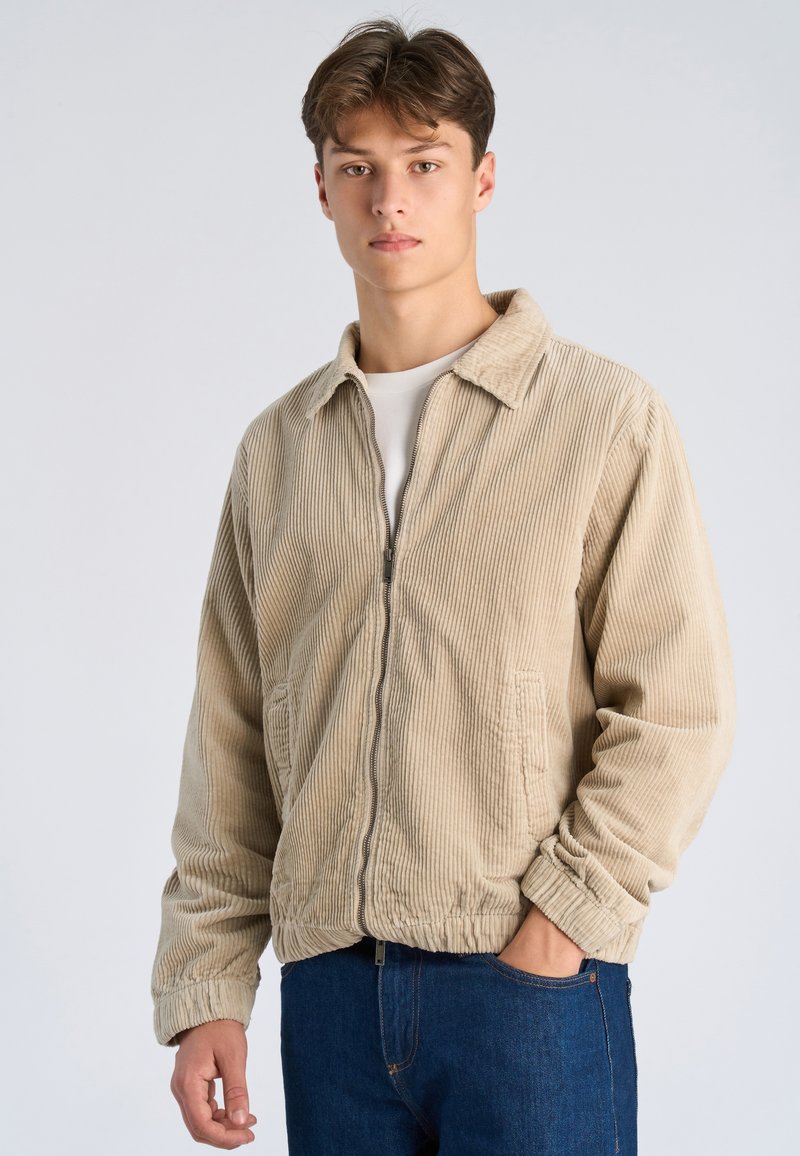 Giovane uomo con capelli castani corti che indossa una giacca di velluto beige sopra una camicia bianca e jeans blu, in piedi davanti a uno sfondo semplice.