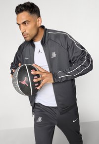 Sort og grå atletisk træningsdragt med hvide accenter, som inkluderer en zip-up jakke og matchende bukser. Holder en sort tekstureret basketball.
