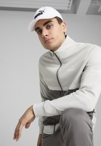 Chaqueta de cremallera gris claro con un panel inferior gris más oscuro, con acentos en blanco. Lleva puesta una gorra blanca con el logo de Puma.