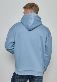 Homme portant un sweat à capuche bleu clair et un jean foncé, de dos, contre un fond gris uni.