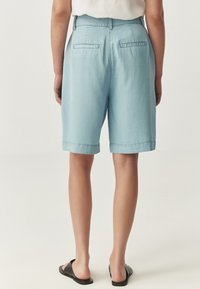 Lichtblauwe shorts van zacht materiaal, met een ontspannen pasvorm, opgerolde randen en twee achterzakken. Draag ze met zwarte slidesandalen.