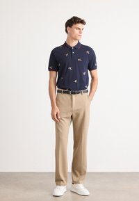 Marineblå polo med flerfargede fuglemønstre, rød kragekant; kombinert med beige bukser og hvite sneakers. Glatt stofftekstur.