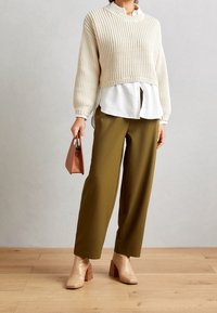 Pull en tricot créme à manches courtes sur une chemise blanche à boutons, pantalon ample olive, chaussures à talons beiges et un petit sac à main marron.