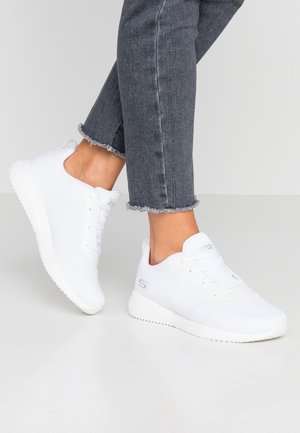 Persoon die witte Skechers-sneakers en versleten grijze skinny jeans draagt, staat op een gladde witte vloer.