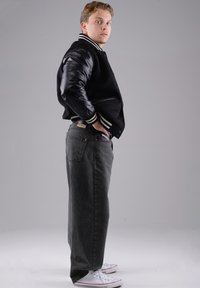 Schwarze Collegejacke mit Lederärmeln, graue lockere Jeans und weiße Sneakers. Die Jacke hat einen gestreiften Kragen und Reißverschlusstaschen.