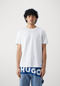 Camiseta blanca de algodón con mangas cortas y cuello redondo, combinada con jeans azules que presentan letras grandes en blanco en los bolsillos, con un tejido texturizado.