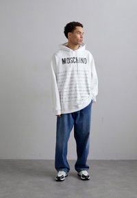 Weißer Kapuzenpullover mit "Moschino"-Logo und sich wiederholendem Textmuster, kombiniert mit hellblauen, trendigen Baggy-Jeans und schwarz-weißen Sneakern.