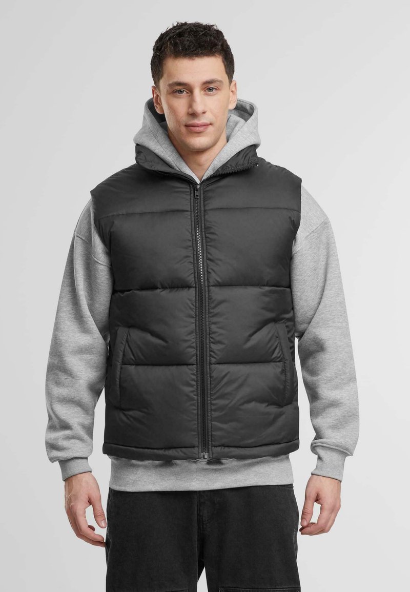 Svart puffervest med glidelås foran og to sidelommer, brukt over en grå hettegenser med snøring. Polstret design med en glattere tekstur.