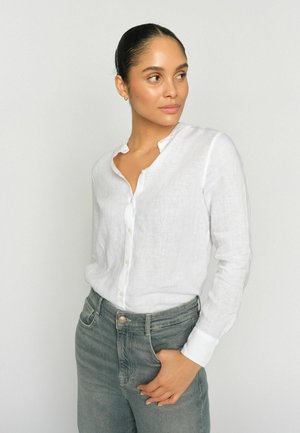 Žena s tmavými vlasmi zviazanými do drdola, oblečená v bielej košeli na gombíky a sivých džínsach, ktorá sa pozerá bokom s jednou rukou vrecku na jednoduchom pozadí.