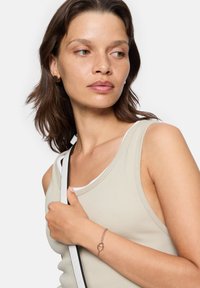 Roséguld armband med ett hjärtformat accent, bärs på en bar handled. Modellen har en neutraltonad linne och håller i en väskarem.