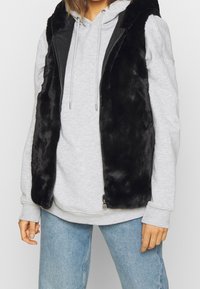 Person som bär en svart fuskpäls ärmlös väst över en ljusgrå hoodie och ljusblå jeans, som står mot en enfärgad bakgrund.