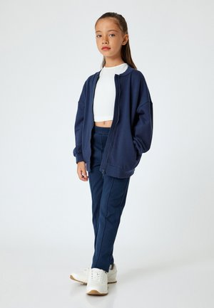 Granatowy, oversize, zapinany na zamek hoodie z miękkiego materiału, w połączeniu z dopasowanymi spodniami i białymi sneakersami. W zestawie znajduje się krótka, biała bluzka.