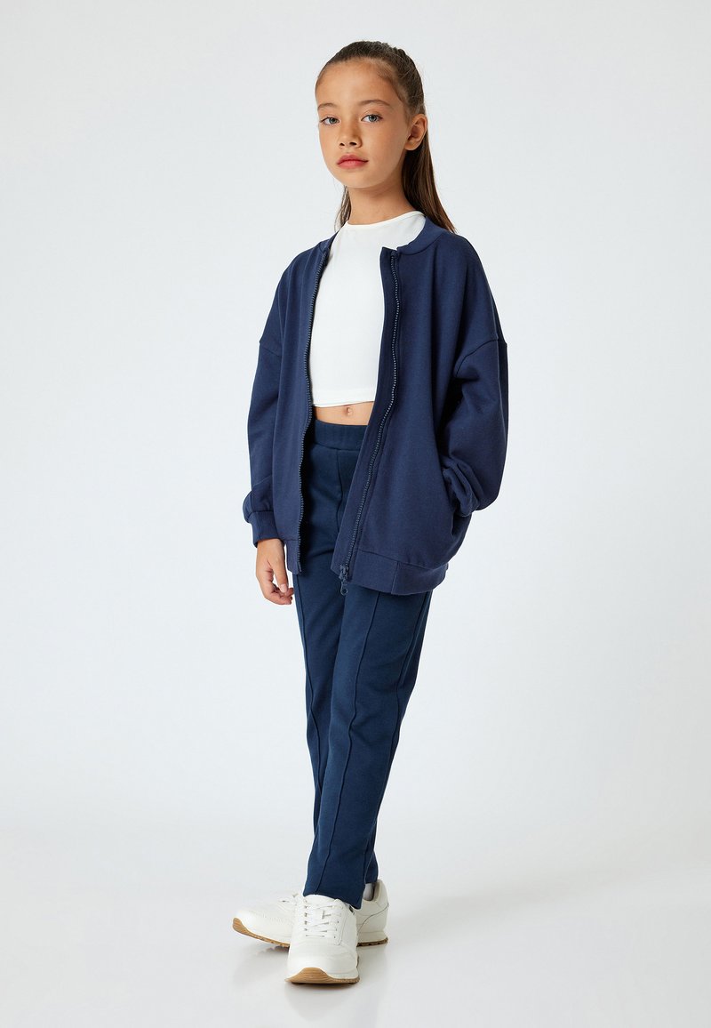 Granatowy, oversize, zapinany na zamek hoodie z miękkiego materiału, w połączeniu z dopasowanymi spodniami i białymi sneakersami. W zestawie znajduje się krótka, biała bluzka.