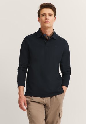 Mann in navy blauem Langarm-Polohemd, beige Cargo-Hosen und einem dunklen Halstuch, stehend mit einer Hand in der Tasche.