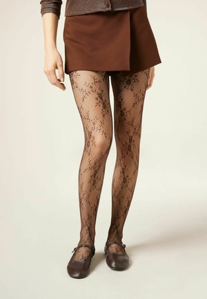 Femme portant une mini-jupe marron, des collants résille noirs à motif floral et des chaussures Mary Jane marron foncé, debout contre un fond uni.