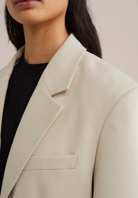 Beige kavaj med en strukturerad krage, en bröstficka och textil med struktur. Bärs över en svart topp, med fokus på slag och axeldetaljer.