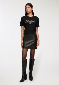 T-shirt nera con la scritta "Delicate", abbinata a una gonna attillata in pelle verde scuro con una fessura laterale, stivali neri fino al ginocchio e collant trasparenti.