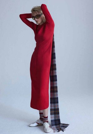 Femme en longue robe rouge avec une écharpe à carreaux suspendue dans son dos, portant des lunettes de soleil, des chaussettes rayées et des baskets blanches, debout les mains sur la tête.