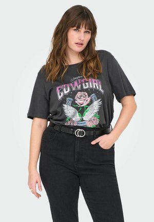 T-shirt graphique noir avec un design de rose et d'ailes, portant le mot "Cowgirl" en lettres majuscules. Associé à un jean noir taille haute et une ceinture.