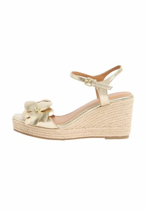 Sandalia de cuña dorada con correa al tobillo y hebilla, plataforma de yute trenzado y una gran decoración de flor dorada en la zona de los dedos.