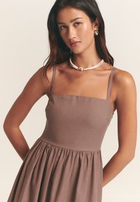 Brun linnklänning med figursydd överdel, tunna axelband och en utsvängd kjol. Accentuera med ett lager-på-lager-halsband och stora runda örhängen.