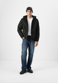 Chaqueta bomber negra acolchada con capucha, cierre de cremallera y puños elásticos; llevada sobre una camiseta blanca y unos vaqueros azules desgastados con zapatos negros.
