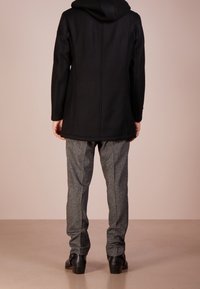 Cappotto nero con cappuccio, dal taglio dritto, abbinato a pantaloni grigi a fantasia e calzature nere. Tessuto liscio, minimalismo nei dettagli.