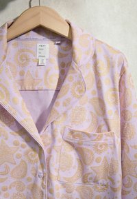 Haut de pyjama lilas avec motifs célestes jaunes et motifs cachemire, devant boutonné, poche poitrine et étiquette indiquant "NEXT time to dream."