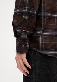 Rechte Hand neben dem dunkelbraun und schwarz karierten Langarmhemd mit Knöpfen, kombiniert mit schwarzen Jeans.