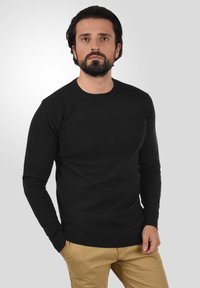 Sort langærmet sweater med et tekstureret strikkemønster, rund halsudskæring og ribbede ærmer og kant. Sammen med brune bukser.