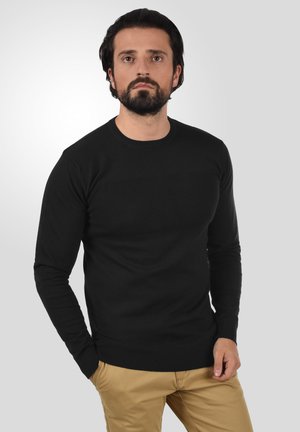 Must soe must pikkade varrukatega sviiter, millel on tekstureeritud kudumismuster, ümar kaelus ja soonikkoes mansetid ning äär. Koos beežide pükstega.