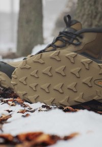 Braune Wanderschuhe mit gemusterten Sohlen, die auf verschneitem Boden mit gefallenem Laub in einer Waldumgebung liegen.