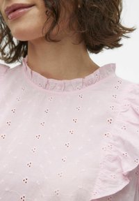 Top rose brodé avec un col et des manches à volants, présentant de petits motifs floraux et un tissu texturé.