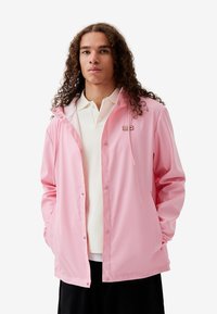 Rosa wasserdichte Jacke mit Kapuze, Druckknopfverschluss und Taschen. Sie verfügt über ein kleines Markenlogo auf der Brust und eine glatte Textur.