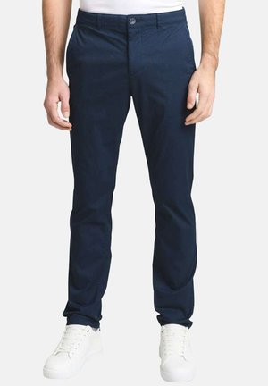 Homme portant un pantalon bleu marine ajusté et des baskets blanches, debout devant un fond clair uni.