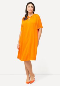 Vestido naranja holgado con escote redondo y mangas cortas, con un detalle de lazo. La modelo lleva unos tacones naranjas a juego.