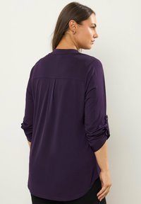 Lila Langarmshirt mit lockerer Passform, mit geknöpften aufgerollten Ärmeln, einem Yoke-Detail auf der Rückseite und einem abgerundeten Saum.