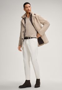 Cappotto in misto lana beige con colletto foderato in pelliccia, maglione beige chiaro a scollo a V, pantaloni bianchi su misura e stivaletti alla caviglia in suede marrone scuro.